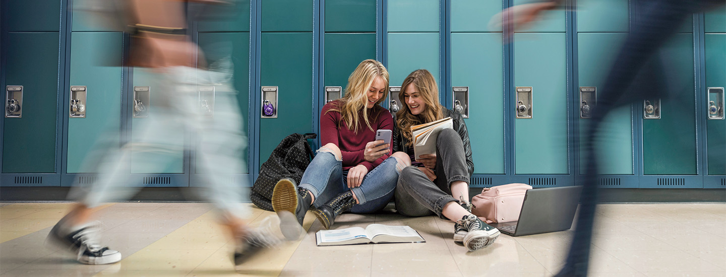 Image of two students looking at a phone while sitting in school hallway